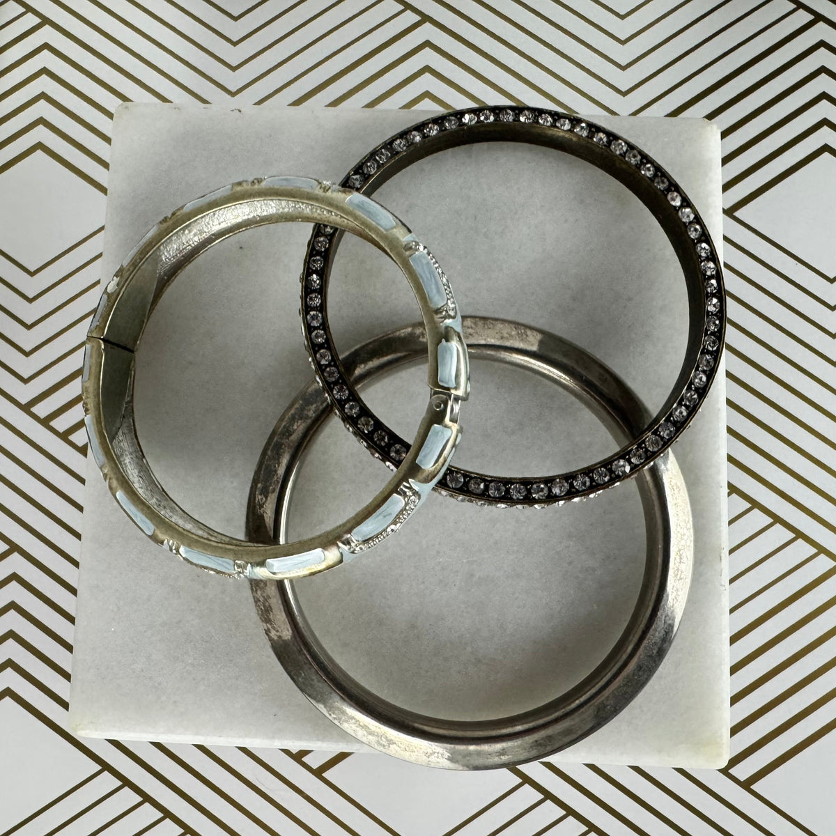 Three silver-toned bangle bracelets arranged on a white square card. Two of the bangles are lined with clear rhinestones, and one bangle features light blue inlay segments.