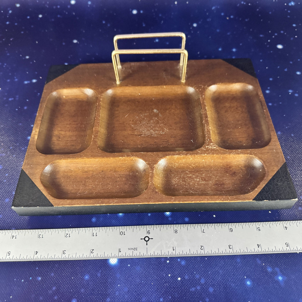 A wooden compartment tray with a gold metal handle, shown next to a metal ruler on a blue starry background.