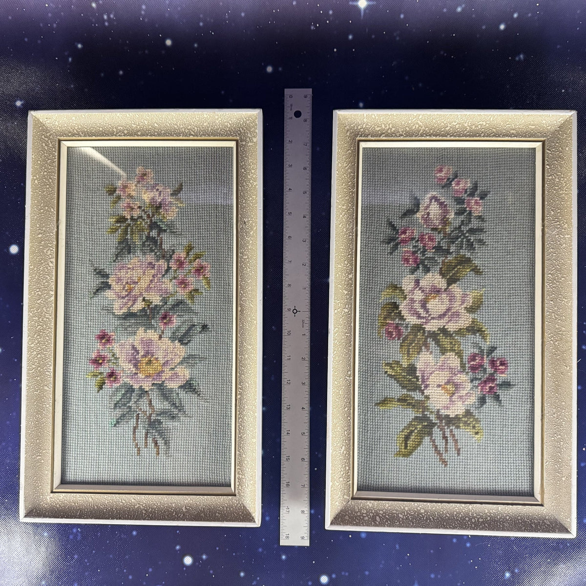 Two framed floral cross-stitch pieces on a dark blue background with a ruler for scale.