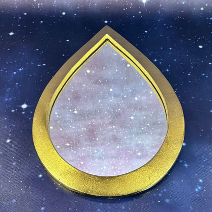 A teardrop-shaped decorative piece with a textured, glittery lavender and pink interior and a thick, polished gold outer frame, set against a dark blue starry background.