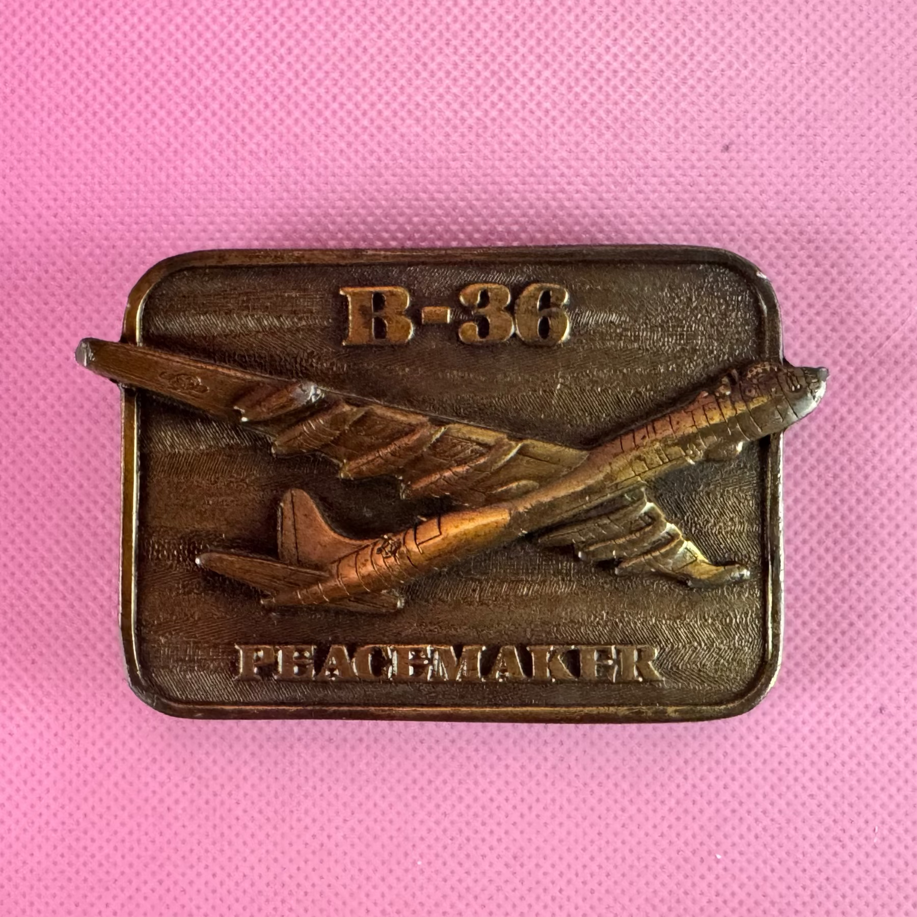 A rectangular bronze-toned metal pin or badge featuring a raised aircraft design with the text 'peacemaker' on the left and 'B-36' on the right, placed on a textured pink background.