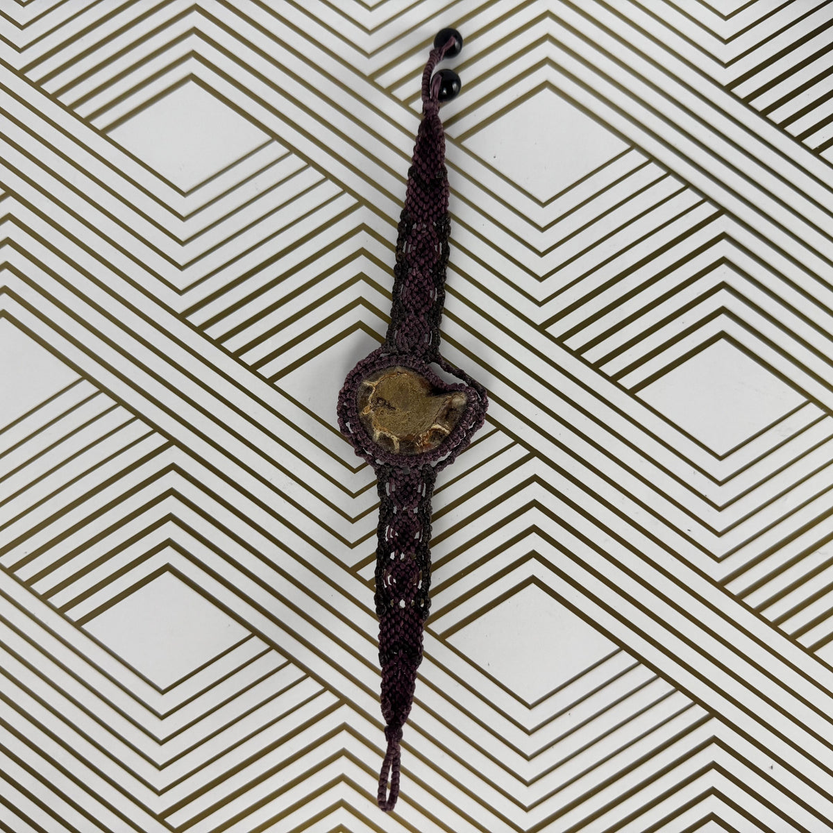 A dark brown braided cord bracelet with a central gold-toned heart-shaped charm, displayed on a white background with a brown geometric pattern.