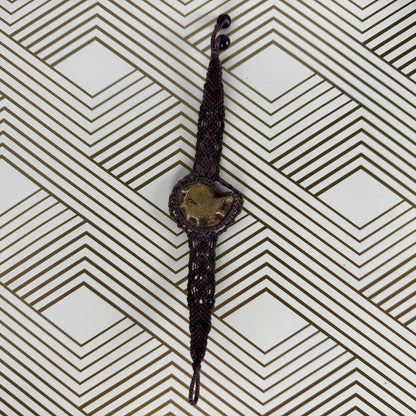 A dark brown braided cord bracelet with a central gold-toned heart-shaped charm, displayed on a white background with a brown geometric pattern.