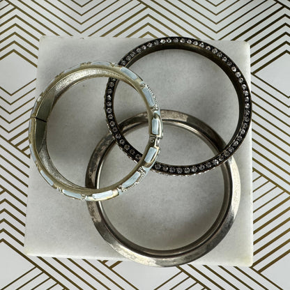 Three silver-toned bangle bracelets arranged on a white square card. Two of the bangles are lined with clear rhinestones, and one bangle features light blue inlay segments.