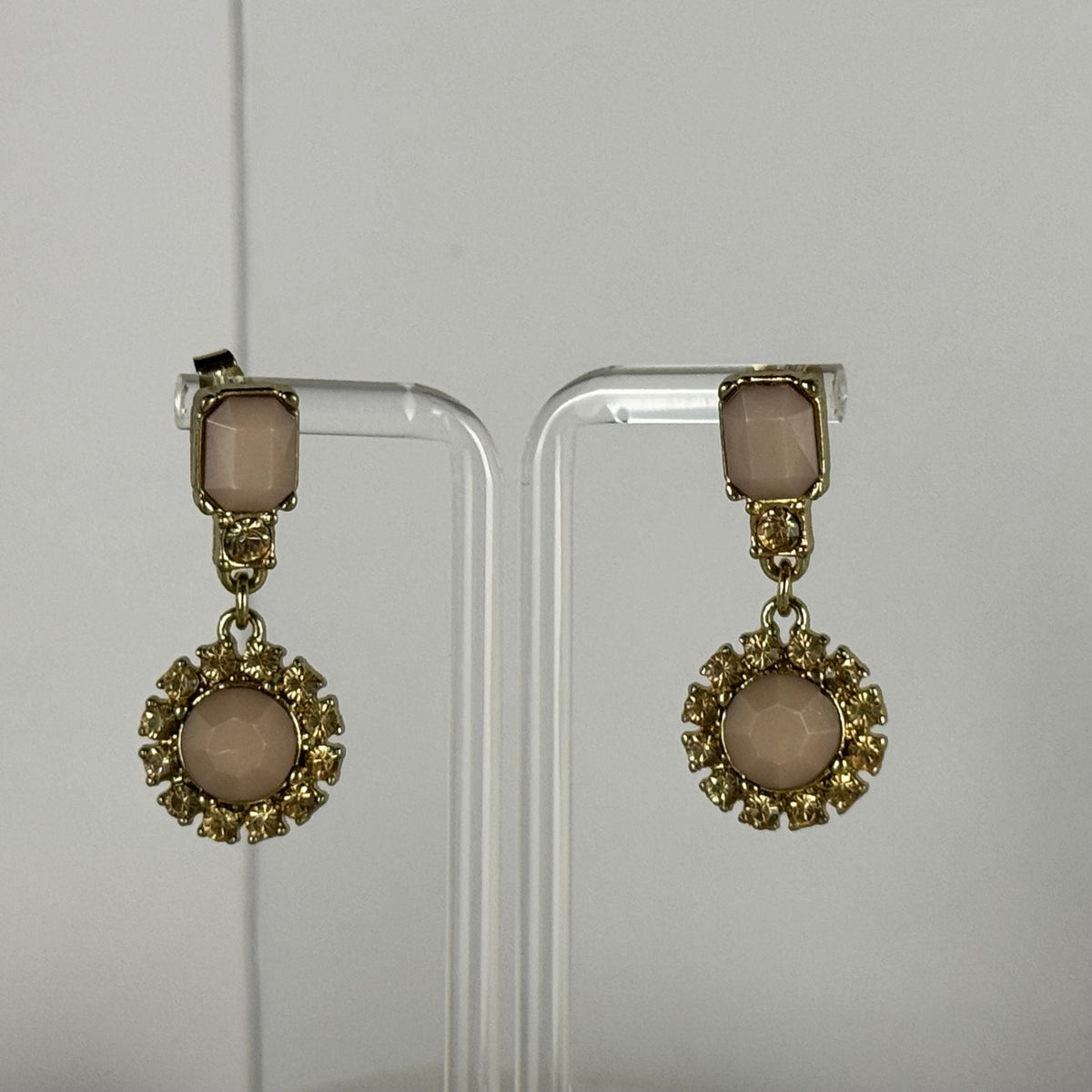 A pair of gold-tone drop earrings with rectangular blush pink stones at the top and round blush pink stones in ornate circular settings below, displayed on clear acrylic stands against a light gray background.