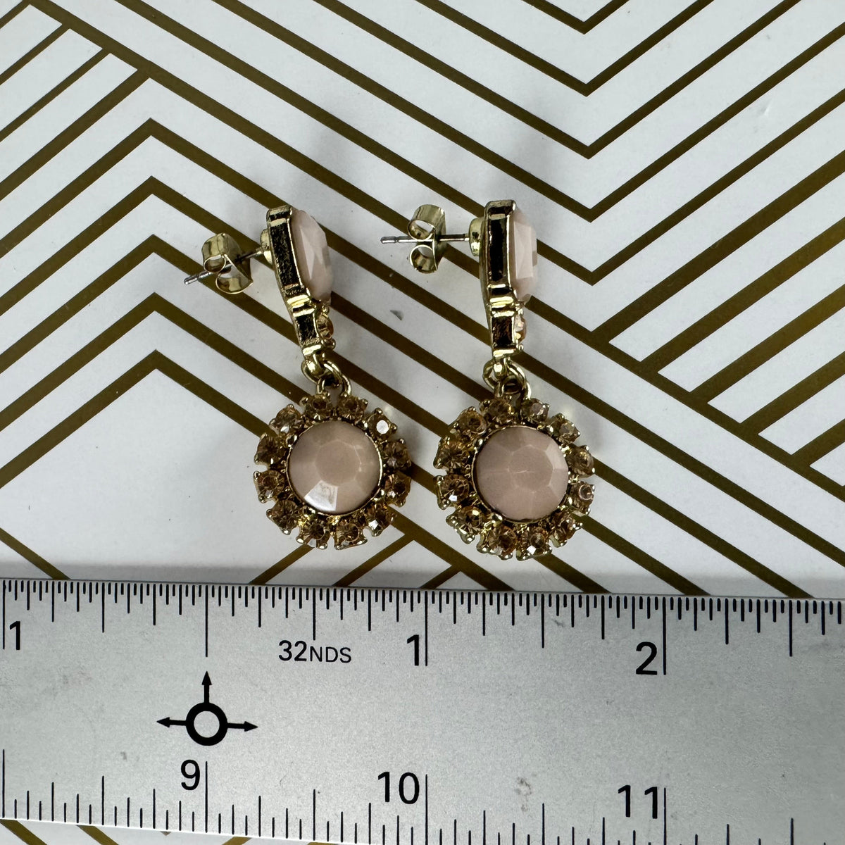 Gold earrings with pink stones on a white background with black chevron pattern and ruler for scale.