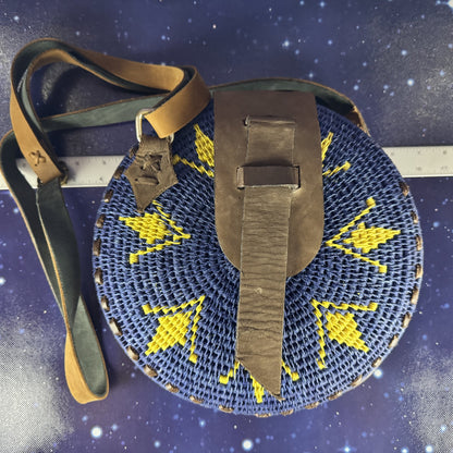 Round blue woven bag with yellow patterns and a brown strap on a dark blue background